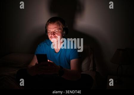 Man wearing headphones using a smartphone in a dark bedroom at night Stock Photo
