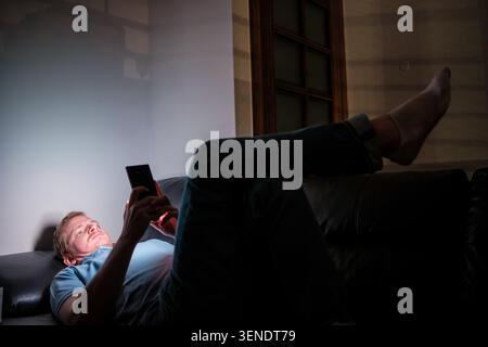 Man lying on sofa, scrolling social media on smartphone in a dark room Stock Photo