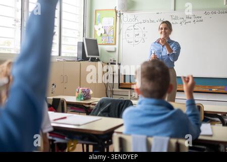 Smiling elementary school teacher leading an interactive English lesson in a bright classroom while students raise their hands to participate. Stock Photo