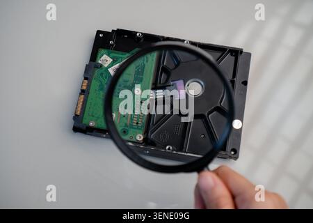 Hand Holding Magnifying Glass Over Disassembled Hard Drive on White Surface Stock Photo