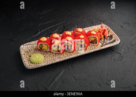 Sushi rolls are placed neatly on a patterned plate. The rolls are topped with red fish roe. Wasabi and pickled ginger are served beside the sushi. The setting has a dark background. Stock Photo