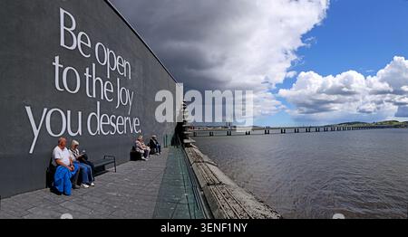 Visitors relaxing outside V&A Dundee beside the Be open to the joy wall, overlooking the River Tay in Scotland Stock Photo