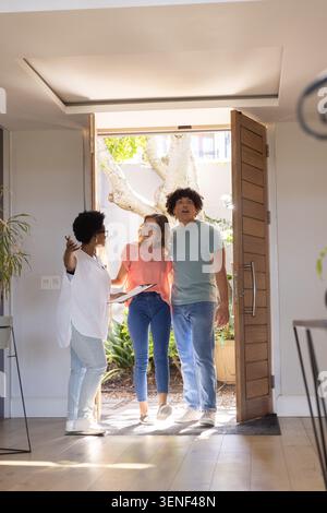 Diverse couple entering home while African American agent gesturing, holding clipboard by open door Stock Photo