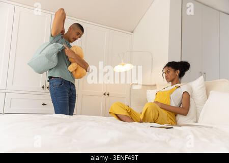Couple man standing holding light-blue bag, tan teddy; woman sitting on bed wearing yellow overalls Stock Photo