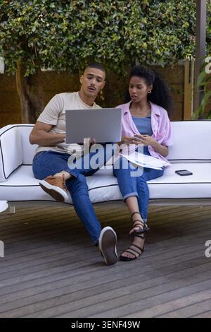 Two colleagues sitting on white sofa outdoors, reviewing laptop and taking notes with pen. Collaboration, teamwork, patio, deck, greenery, daylight, c Stock Photo