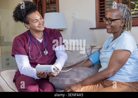 Caregiver in scrubs and Senior African American woman sitting on sofa checking BP using monitor Stock Photo
