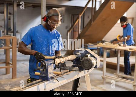African American coworkers in blue T-shirts operating wood lathe and measuring wooden plank in shop Stock Photo