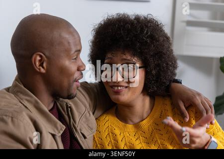 Couple sitting on couch at home, man wearing jacket and watch, woman wearing eyeglasses and sweater Stock Photo