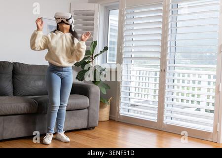 Child girl standing using VR headset in living room wearing cream hoodie, blue jeans, copy space Stock Photo