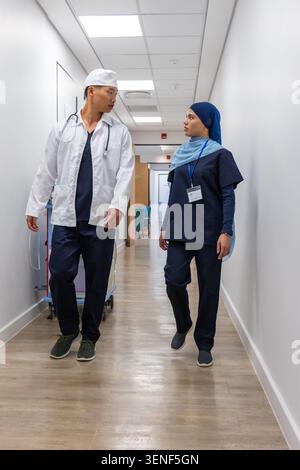 Diverse coworkers walking and talking in hospital corridor wearing navy scrubs stethoscope ID badge Stock Photo