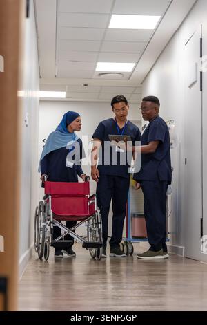 Diverse medical team checking tablet beside red wheelchair in clinic hall, wearing navy scrubs IDs Stock Photo