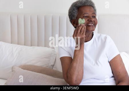 Senior African American woman sitting on bed in white top by headboard using heart guasha smiling Stock Photo