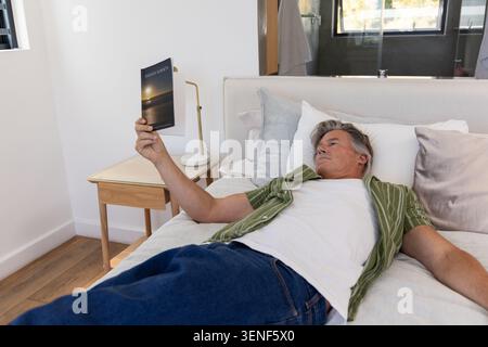 Middle-aged man lying on bed in bedroom holding paperback book and wearing green shirt Stock Photo