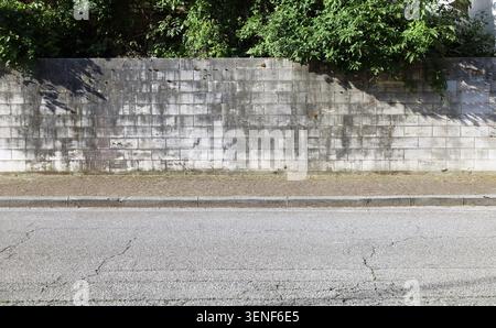 Weathered surrounding wall made of stone tiles. Trees on behind, sidewalk and street in front. Grunge background for copy space. Stock Photo