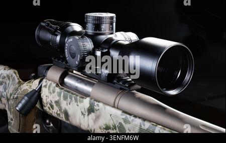 Bolt action rifle with a camouflage paint job with a large magnified rifle scope on top with a dark background behind. Stock Photo