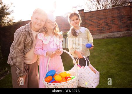 Three kids pose with baskets of colorful Easter eggs in a backyard, wearing festive clothing, with a sunny atmosphere and greenery in the background Stock Photo