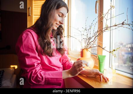 Young woman uses a skincare product on her hand near a window, with decorative plants and skincare items placed on the windowsill Stock Photo