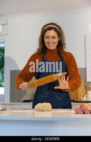 Woman wearing sweater, denim apron standing at kitchen island holding rolling pin, preparing dough Stock Photo