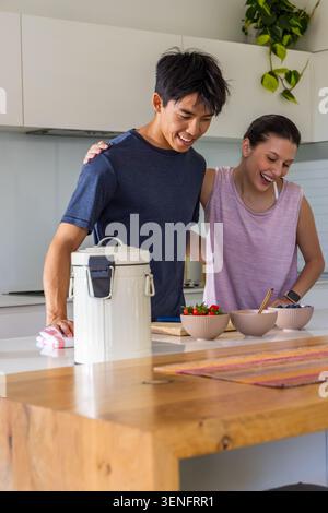 Diverse couple preparing three ceramic bowls at kitchen island, wearing navy t-shirt and pink tank Stock Photo