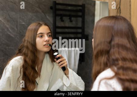 Young Woman Brushing Teeth with Electric Toothbrush. Young woman in a white bathrobe brushes her teeth with a black electric toothbrush while looking Stock Photo