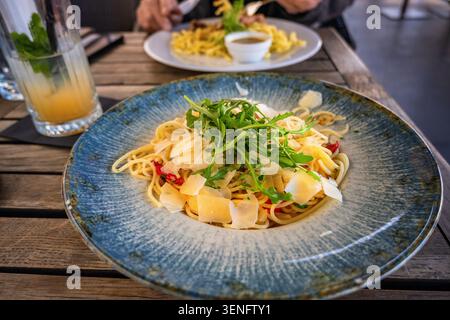 Spaghetti aglio olio pepperoncino with parmesan and arugula leaf on blue plate, lemonade and second meal on restaurant table. Stock Photo