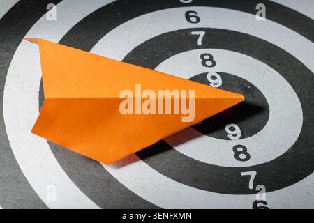 A close-up top-down view showing an orange folded origami paper airplane placed on a black and white numbered dartboard, symbolizing modern innovation, market disruption, and hitting strategic destinations. Stock Photo