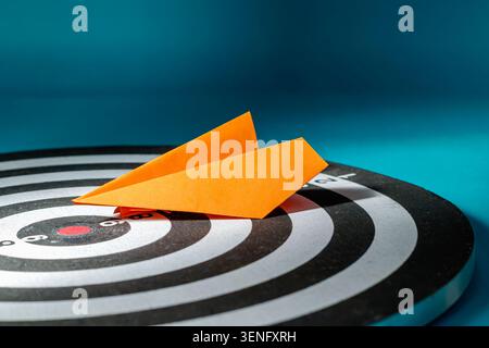 A low-angle close-up shot of an orange folded paper airplane resting on a circular dartboard, directed toward the target center, symbolizing a creative approach, direct marketing, and innovative business goals. Stock Photo