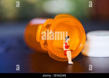 Miniature woman in red dress sits inside an orange pill bottle on a dark surface, with a white cap beside it and a blurred green background Stock Photo