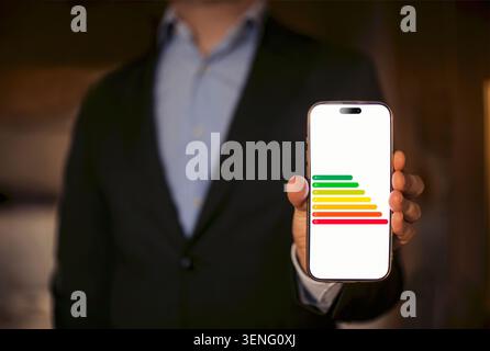 Businessman in a black suit presents smartphone with energy efficiency graphic, showcasing colored bars indicating performance levels in a modern offi Stock Photo