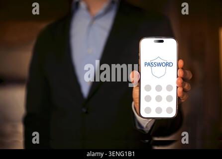 Businessman in a black suit presents a smartphone with a password entry screen, set against a blurred background, highlighting mobile security and tec Stock Photo