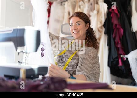 Sewing clothes in a workshop with a smiling woman working on a sewing machine and various fabrics around her Stock Photo
