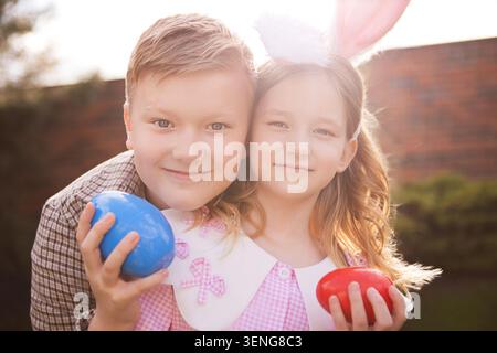 Two kids, a boy and a girl, pose with blue and red Easter eggs in a sunny outdoor environment, surrounded by greenery and a brick wall Stock Photo