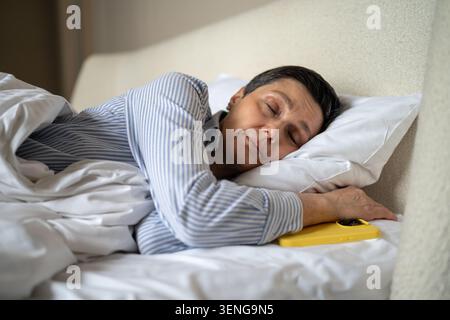 Calm senior woman comfortably sleeping in bed in at home with smartphone lying beside pillow. Stock Photo
