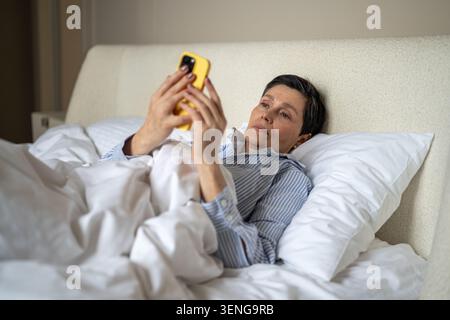Relaxed senior woman scrolling media feeds online on smartphone while lying in bed in bedroom. Stock Photo