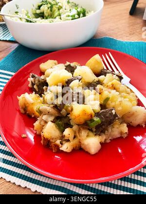 A wooden table holds a bright red plate filled with potato and beef stew next to a large bowl of fresh salad. This meal offers a nutritious option for Stock Photo