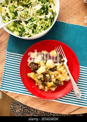A wooden table holds a bright red plate filled with potato and beef stew next to a large bowl of fresh salad. This meal offers a nutritious option for Stock Photo