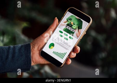A person holds a smartphone showing a healthy eating guide with green visuals and charts, set against a softly blurred background of plants Stock Photo