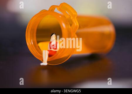 Miniature woman in a red dress is seated inside an orange pill bottle, with a dark blurred background creating a striking visual contrast Stock Photo