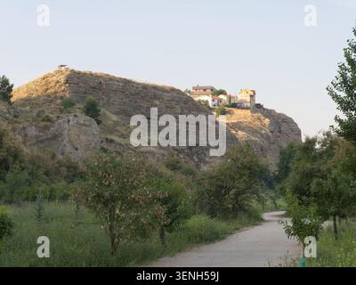 A path runs through green fields leading to a village on a hilltop. The sun shines down on the rocky terrain and trees along the path. People may walk Stock Photo