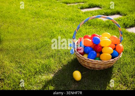 Wicker basket filled with vibrant plastic Easter eggs sits on lush green grass, with one egg placed on the ground beside it Stock Photo