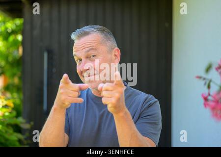 Cheerful mature man pointing at viewer with playful finger gesture and confident smile outdoors. Authentic lifestyle portrait representing communicati Stock Photo