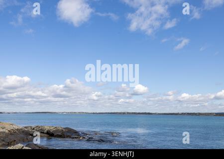 Peaceful coastal scene with rocky shoreline and calm blue water beneath a wide blue sky filled with scattered white clouds overlooking a distant Canad Stock Photo