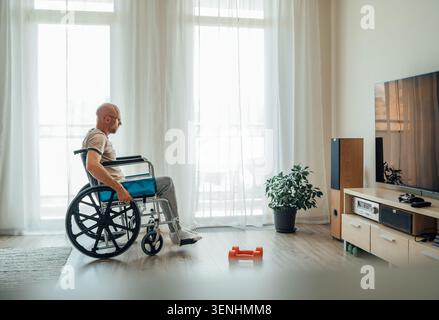 side view of man in wheelchair sitting in bright living room looking at orange dumbbells on floor nearby representing adaptive fitness physical rehabi Stock Photo