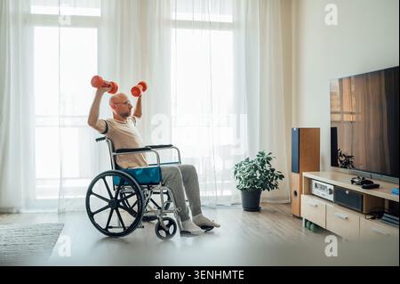 side view of active man in wheelchair doing overhead shoulder press exercise with red dumbbells in bright living room near tv representing adaptive fi Stock Photo
