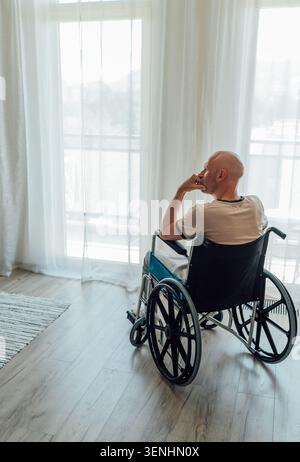 thoughtful man in wheelchair sitting alone in bright room looking out window representing reflection physical rehabilitation journey and hope for futu Stock Photo