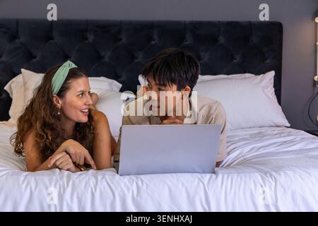 Diverse couple lying on bed in bedroom, propping on elbows, wearing green headband and using laptop Stock Photo
