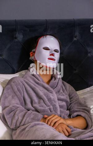 Asian man reclining on bed against tufted headboard, wearing white LED mask and gray robe Stock Photo