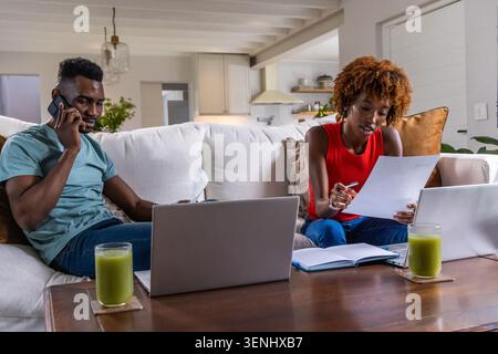 African American couple sitting on light-beige sofa at home, using laptops, phone and paperwork Stock Photo