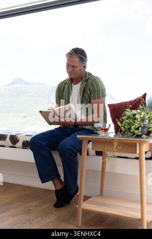 Mature man sitting on window bench at home, wearing green shirt, writing with pen in notebook Stock Photo