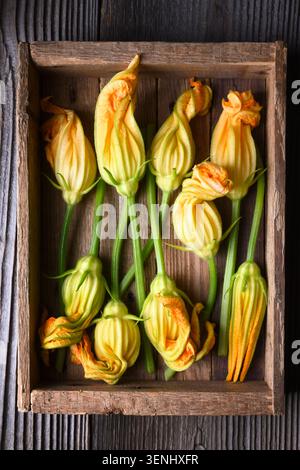 Overhead zucchini with blossoms arranged in wooden box on dark wooden surface. Fresh organic vegetables with edible flowers and rustic styling Stock Photo